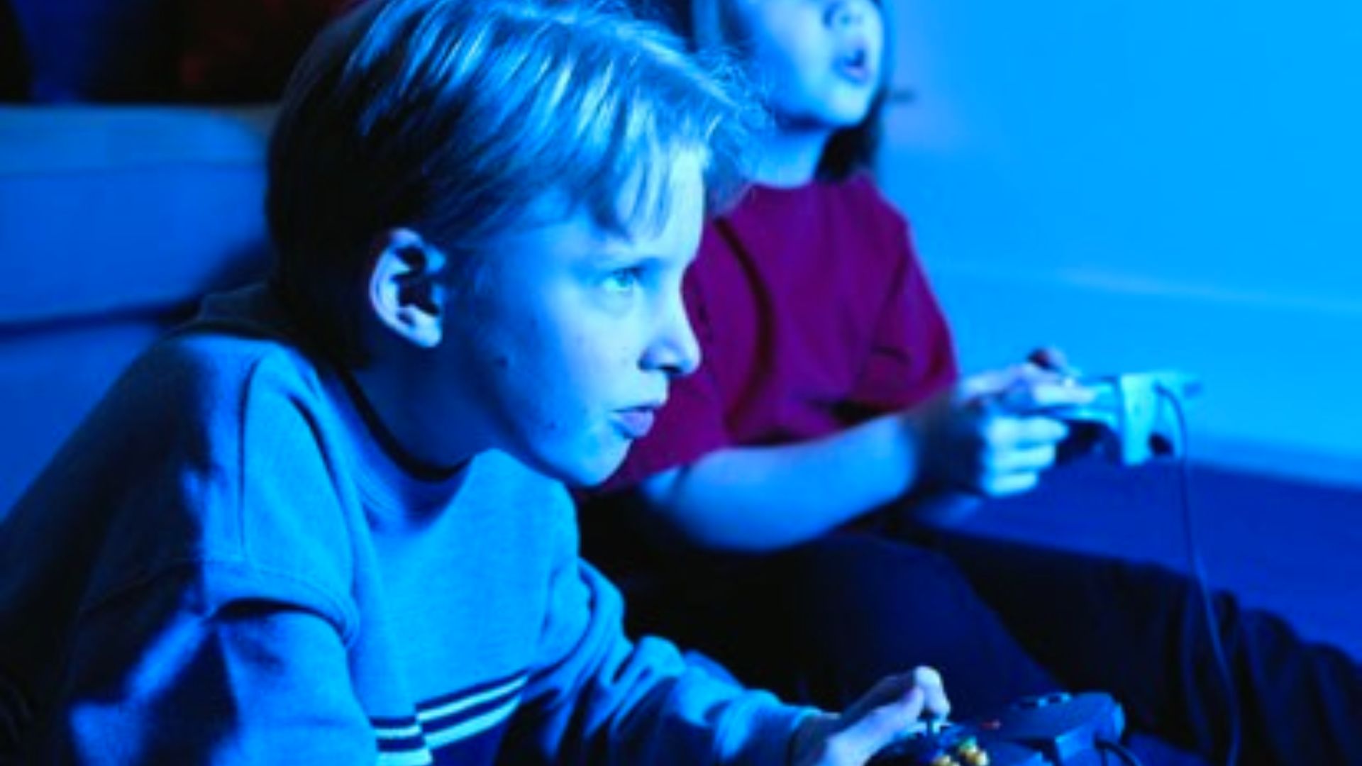 "Two children playing video games in a dark room lit by blue light from the screen. A boy in the foreground wears a dark shirt with stripes and focuses intently on his controller. A child in a red shirt sits slightly behind him."