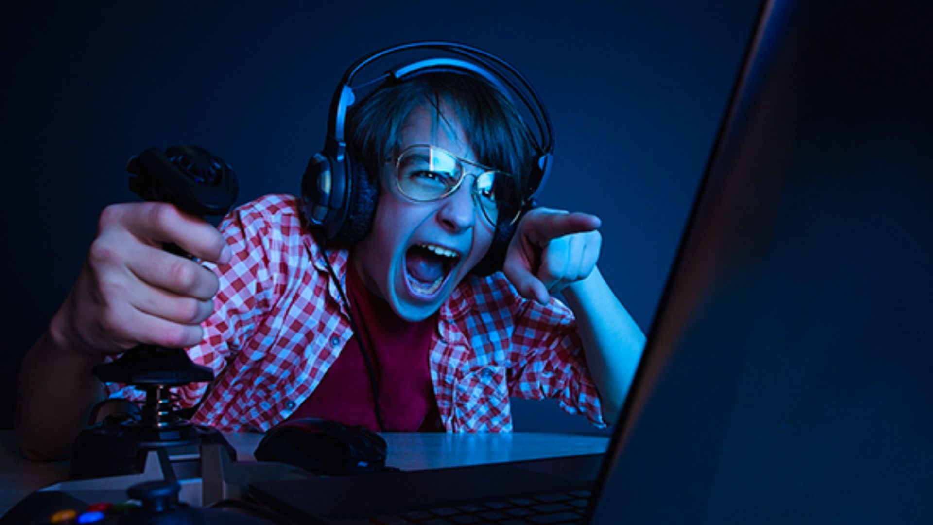 "A young person in a checkered shirt and glasses passionately plays a computer game. They hold a joystick and wear headphones, with an excited expression."