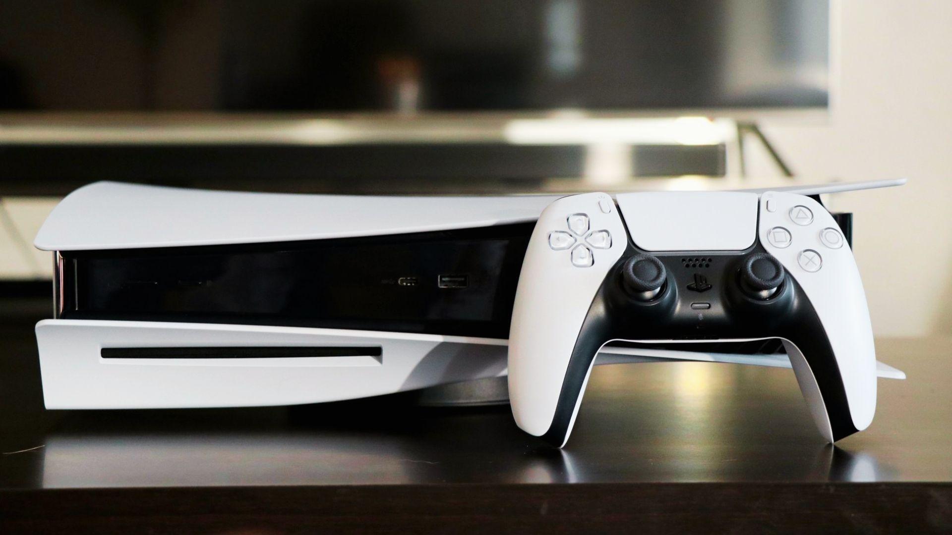 White PlayStation 5 console with disc drive and DualSense controller on dark table, blurred TV and soundbar background
