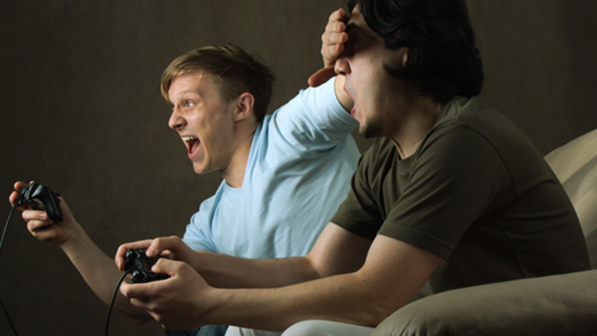 "Two men sitting on a couch playing video games. The man on the left in a light blue shirt yells with his mouth open. The man on the right in a dark green shirt covers his face with one hand while holding a controller in the other."