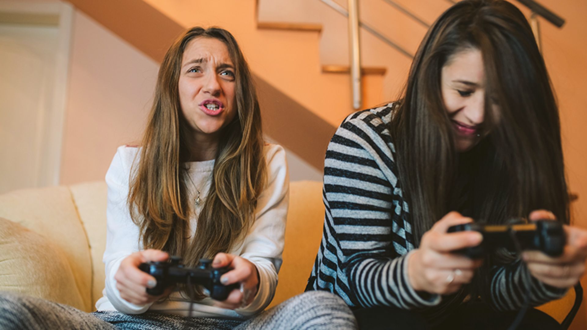 "Two young women sitting on a light-colored couch playing video games. The woman on the left in a white top looks frustrated and is shouting, while the woman on the right in a black-and-white striped shirt smiles and looks down at her controller."