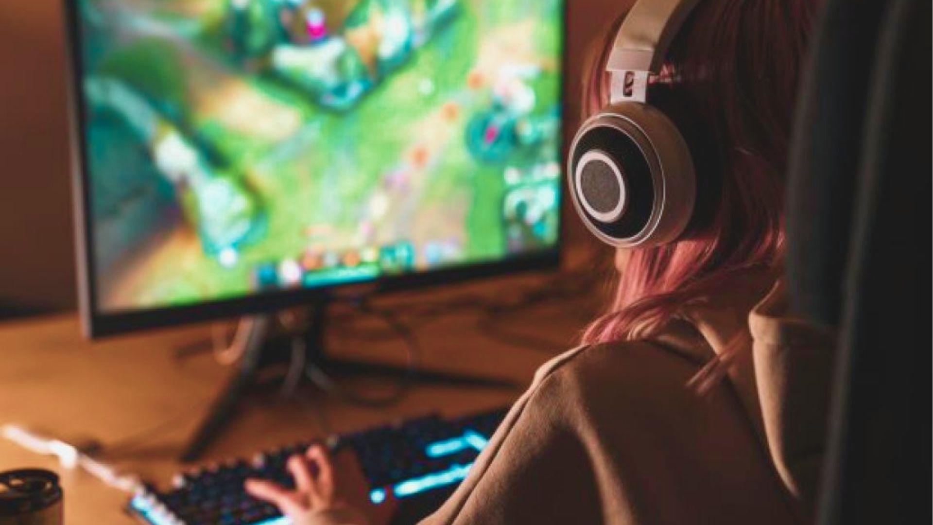 A gamer with pink hair wearing headphones focuses on a strategy game on her monitor.