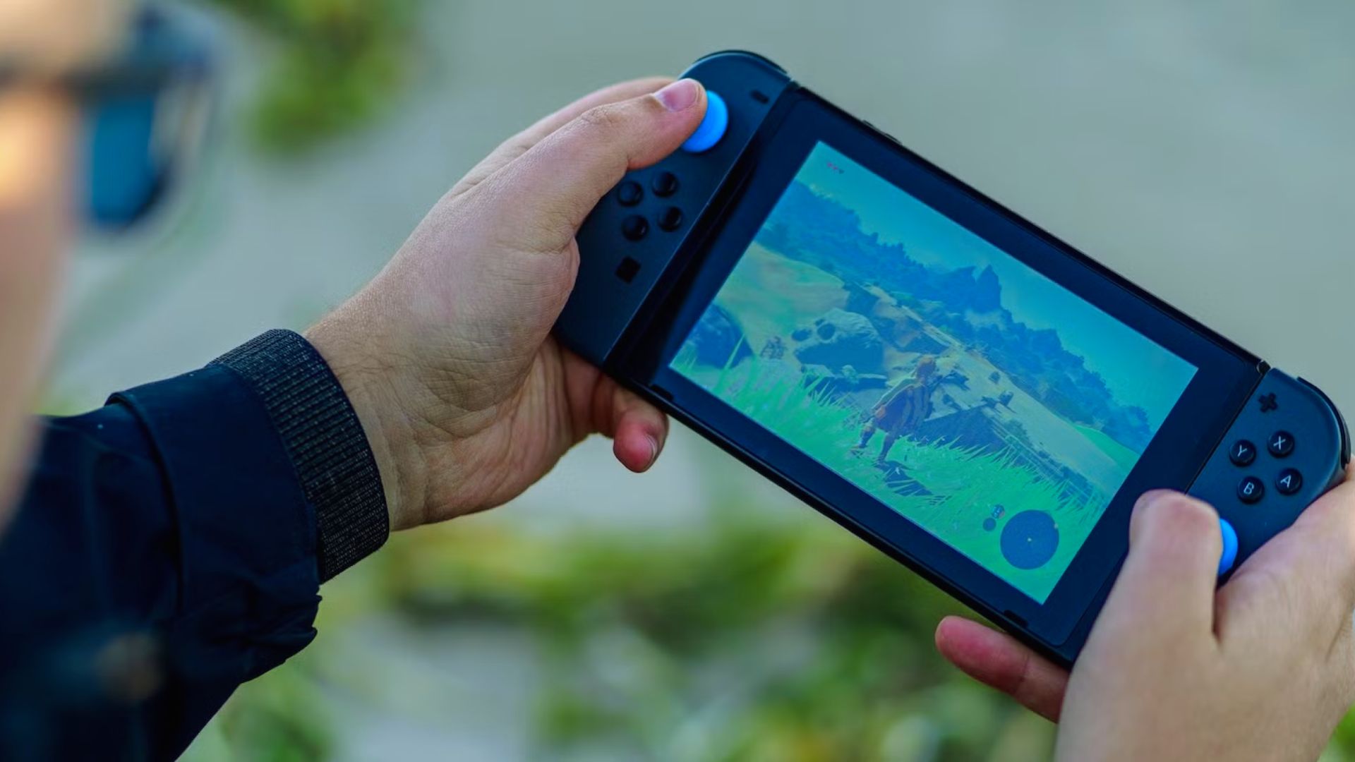 "Close-up of a person's hands holding a handheld gaming console outdoors. The console has dark blue controllers with light blue joysticks. The screen displays a colorful open-world adventure game showing a character standing in tall green grass with mountains in the distance. The background is softly blurred, suggesting a park or natural setting."