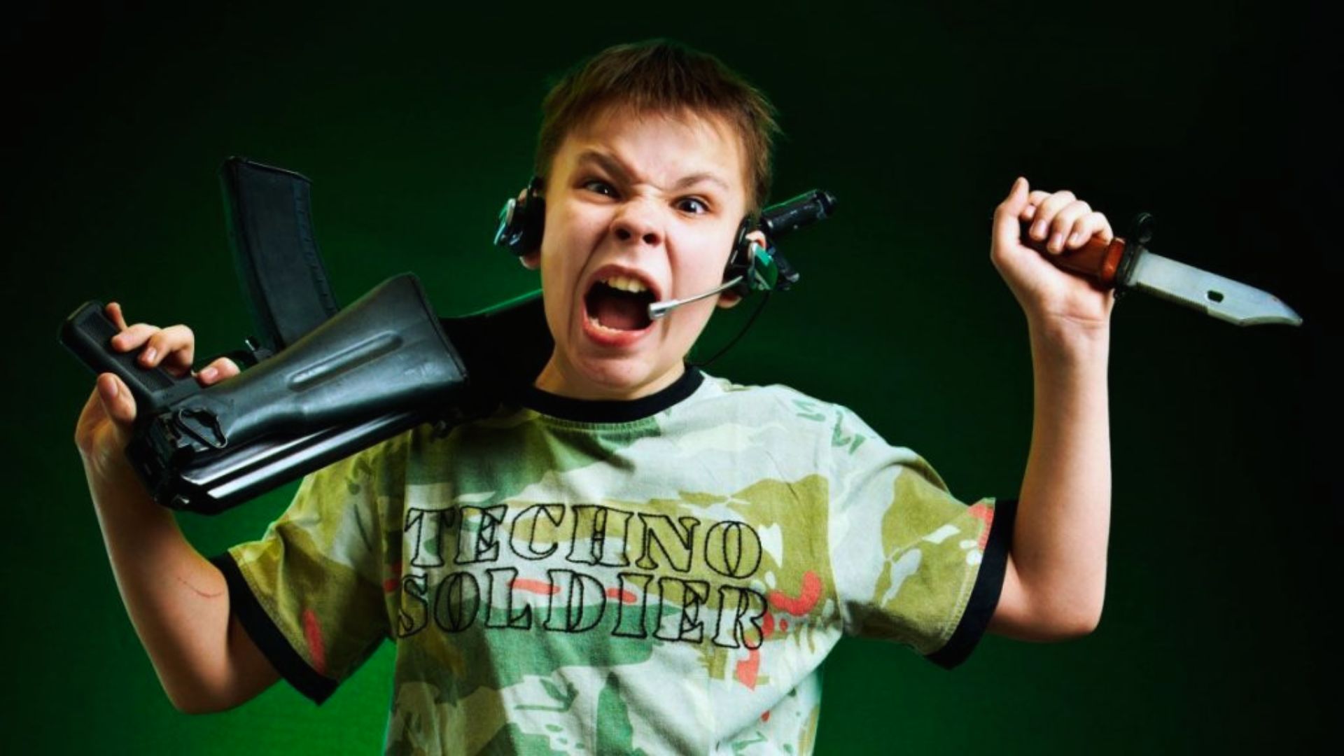 "A young boy wearing a headset and a camouflage "Techno Soldier" t-shirt shouts while holding a toy rifle over one shoulder and a knife in the other hand. The background is dark green."