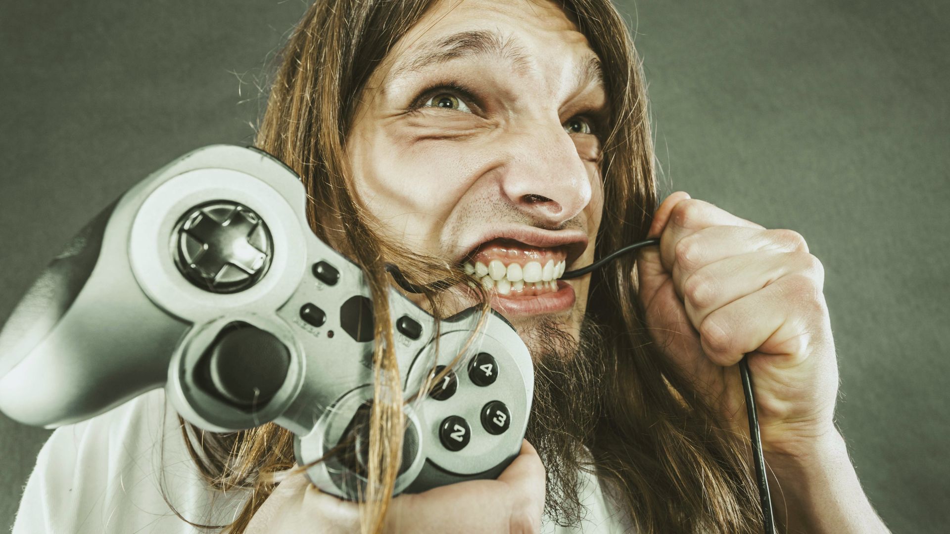 A man with long hair and a beard angrily bites the cord of a video game controller while holding the controller in his other hand. His expression shows frustration.