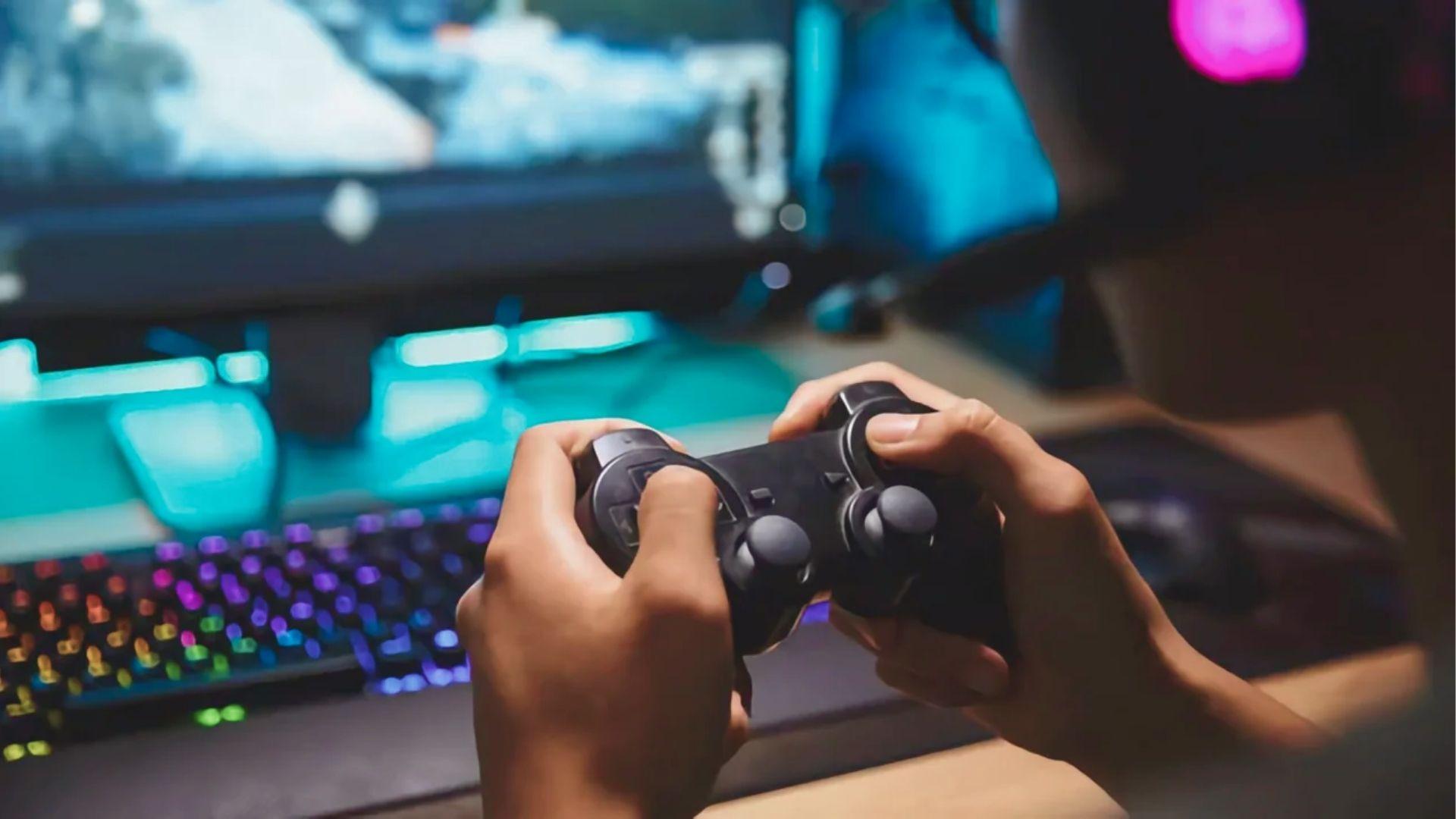 A gamer grips a console controller while an RGB keyboard lights up the desk behind them.