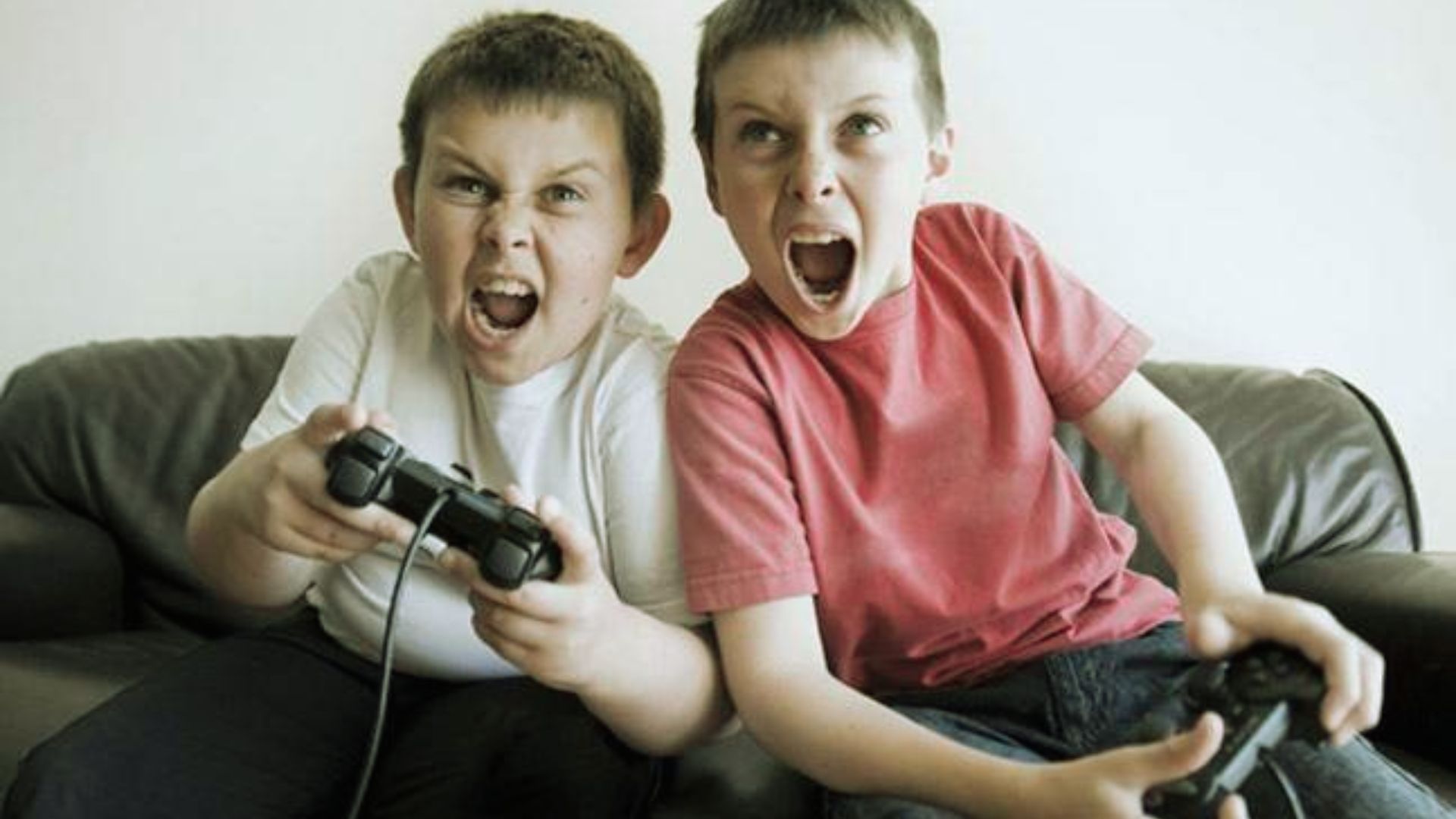 Two young boys sitting on a gray couch, yelling with intense expressions while holding video game controllers. One wears a white t-shirt and the other a red t-shirt."