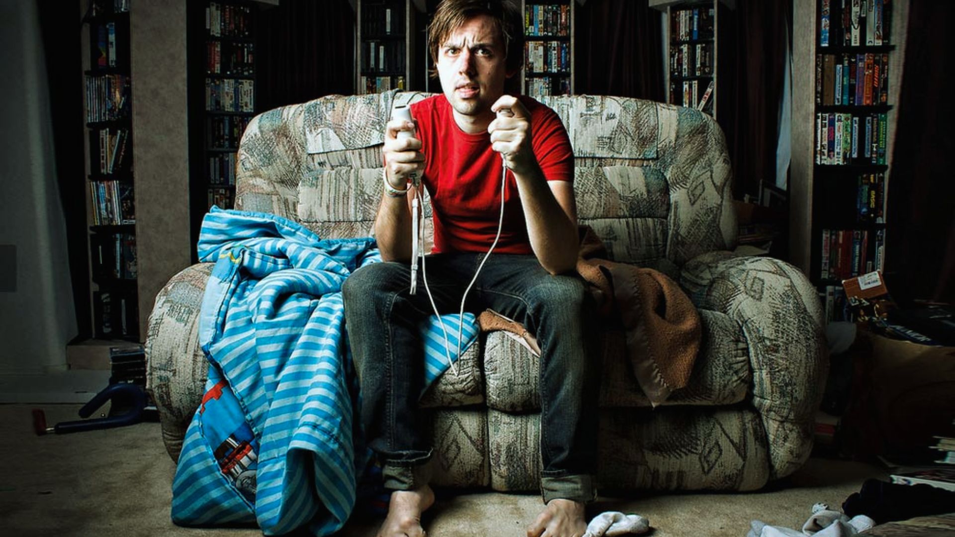 "Man intensely playing video games on a patterned couch, surrounded by clutter